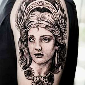 lackwork realism tattoo on an upper arm featuring a woman's portrait with a winged headdress and sunflowers.