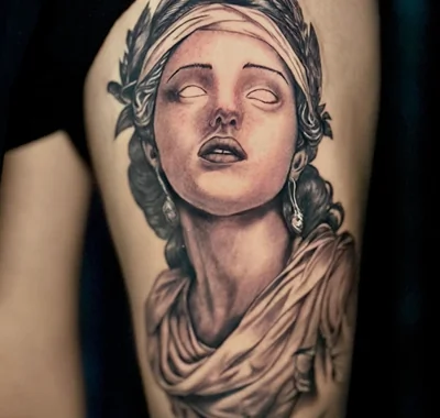 Large detailed black and grey portrait tattoo of a classical goddess statue with draped fabric on a leg.
