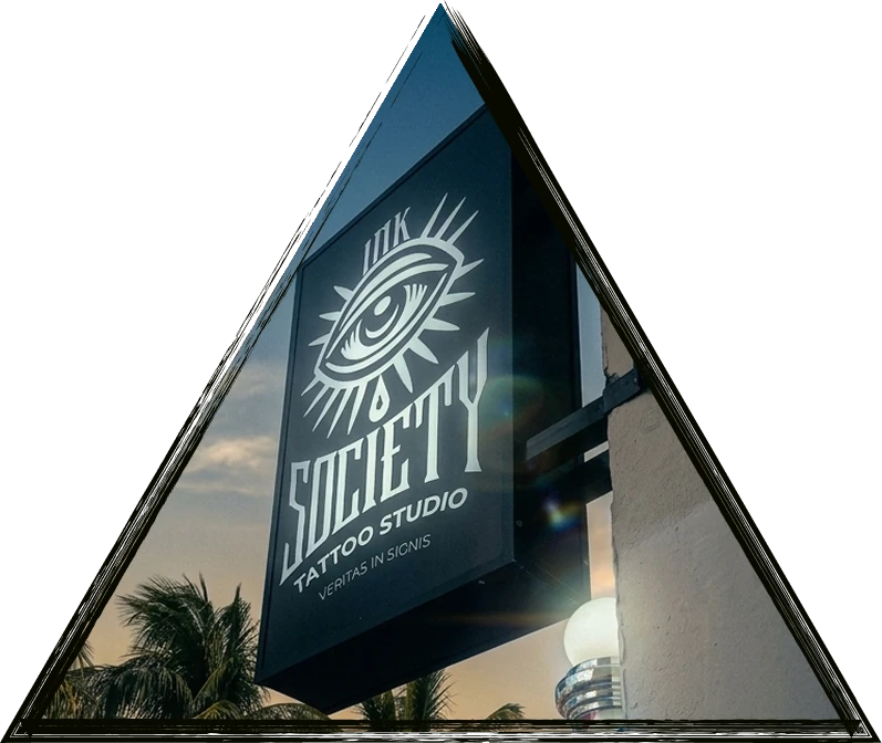 Black exterior hanging sign for a professional tattoo studio featuring a stylized eye logo, mounted on a building wall with palm trees and a sunset sky in the background.