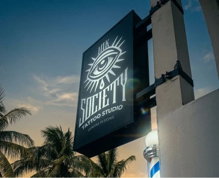 Black outdoor hanging sign for Ink Society Tattoo Studio featuring a stylized eye logo, set against a warm tropical sunset sky with palm trees.