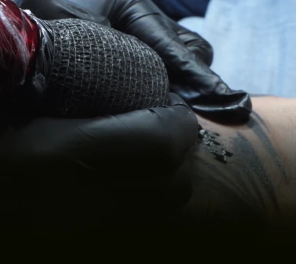 Close-up of a tattoo artist's hands in black gloves using a wrapped tattoo machine to ink a design on a client's skin.