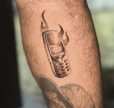 Custom black and grey small tattoo featuring a retro brick-style cell phone engulfed in flames, created with precise dotwork and stipple shading on a person's arm.