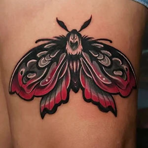 Tattoo of a stylized moth on skin, featuring deep black and vibrant red color blending on its wings with intricate dark patterning.