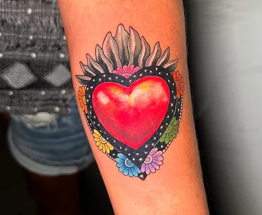 Vibrant full-color custom tattoo on an arm, featuring a detailed glowing sacred heart with flames, surrounded by colorful flowers.