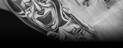 Black and grey tattoo on an arm depicting expressive comedy and tragedy theater masks. Click to contact us for an appointment.