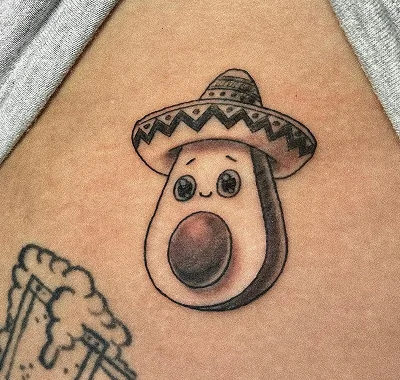 Small, cute custom tattoo of a smiling avocado with large expressive eyes wearing a traditional woven sombrero, featuring soft black and grey shading on a person's skin.