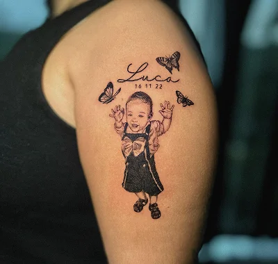 Realistic medium black and grey portrait tattoo of a smiling toddler surrounded by delicate butterflies and lettering.
