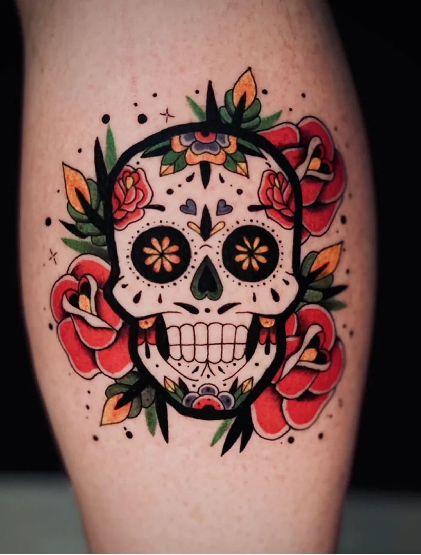 A vibrant neo-traditional style tattoo of a Mexican sugar skull (calavera) on a person's leg. The skull features intricate floral patterns inside the eye sockets, heart shapes, and colorful forehead decorations, framed by large red roses, green leaves, and black stippling dots.