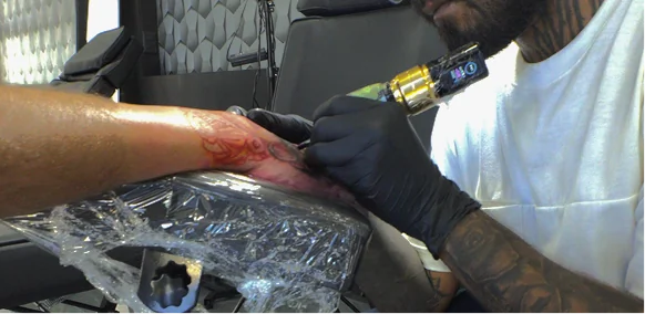Professional tattoo artist wearing black gloves using a modern rotary tattoo machine to carefully ink a custom design on a client's hand during a session.