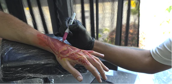 Professional tattoo artist wearing black gloves drawing a custom freehand design with a red marker on a client's hand over a sterile, plastic-wrapped armrest.