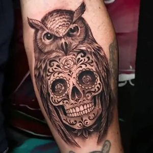 Blackwork realism tattoo on a limb featuring an owl perched above a decorated sugar skull with roses in its eye sockets.