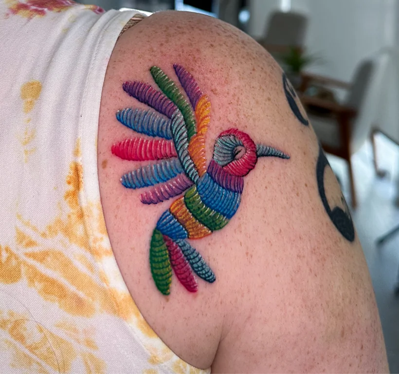 Vibrant color tattoo on a shoulder depicting a hummingbird, designed in a realistic embroidery patch style with textured, rainbow-colored thread lines.