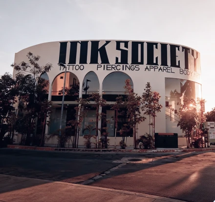 Exterior view of a professional tattoo, piercing, and body art studio building during sunset, featuring large arched windows and a tropical street lined with palm trees.
