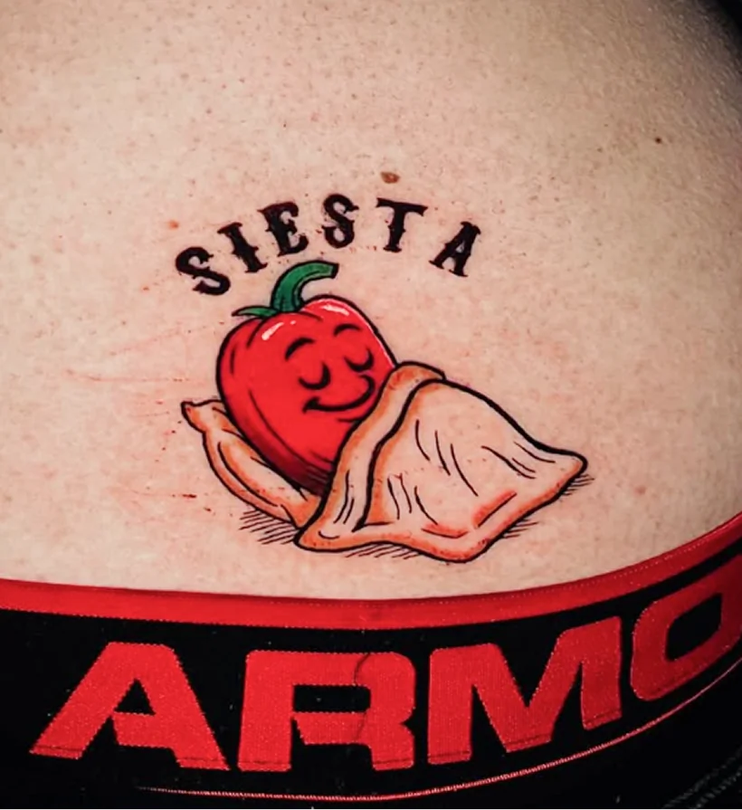 Small color tattoo of a smiling red pepper sleeping tucked under a blanket, with the word "SIESTA" arched above it in black lettering