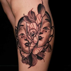 Blackwork tattoo on a thigh featuring a split Japanese mask revealing a large blooming flower in the center.