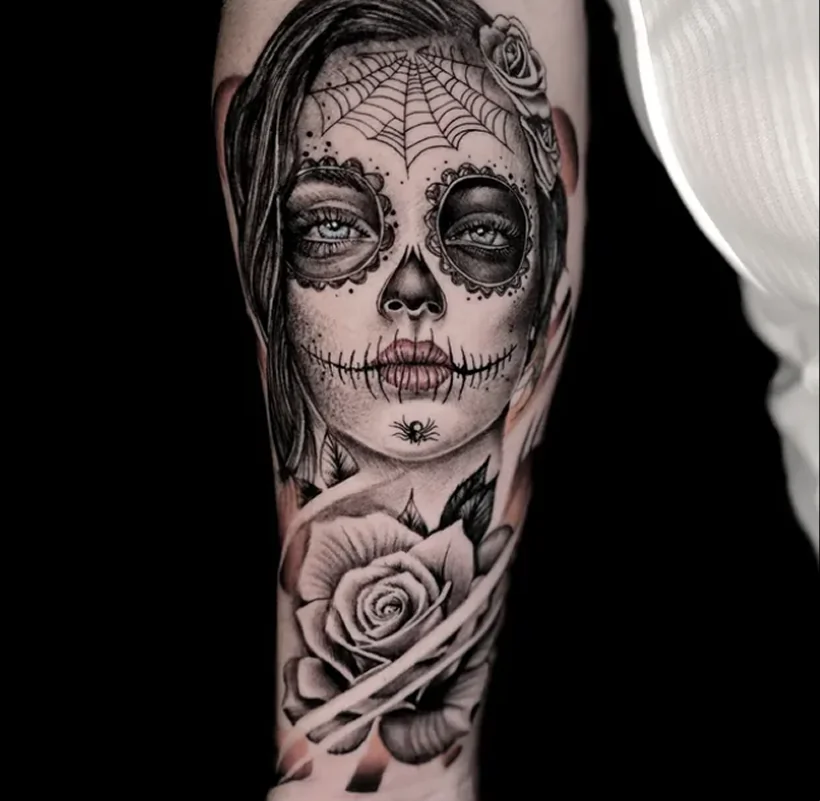 Black and grey realism tattoo on a forearm, depicting a woman with Day of the Dead (Catrina) skull makeup, a spiderweb on her forehead, and a large rose below.