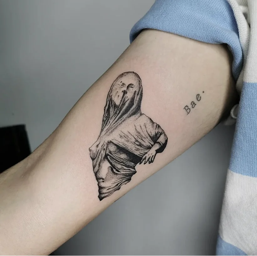 Black ink fine line tattoo on a forearm, depicting a figure entirely draped in cloth with only the arms showing, alongside the word 'Bae.'