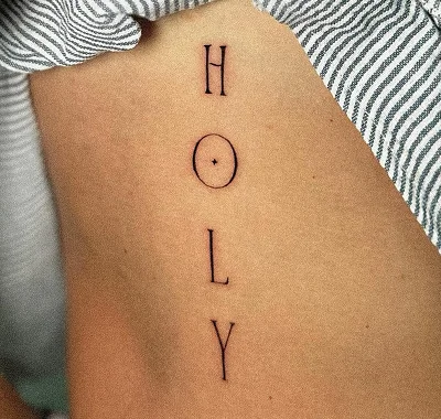 Minimalist fine line lettering tattoo featuring the word Holy written vertically in a delicate, thin serif font on a person's ribcage.