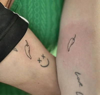 Minimalist fine line matching tattoos featuring small, delicate chili peppers on the inner forearms of two people resting side-by-side.