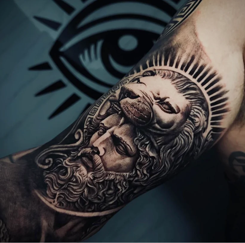 Black and grey realism tattoo on an arm, featuring a classical bearded face wearing a lion headdress, surrounded by a sunburst design.