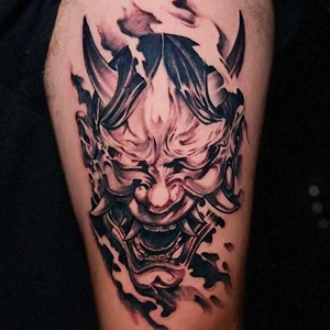 Black and grey tattoo on a limb featuring a Japanese Hannya mask with horns, sharp teeth, and dark smoky shading.