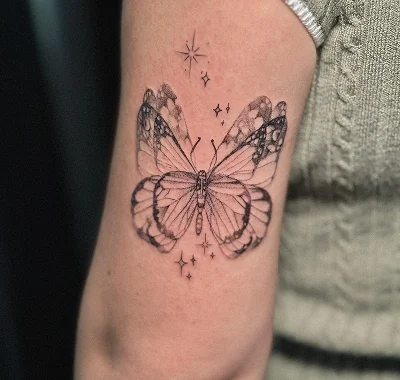 Detailed small black and grey fine line tattoo of a beautiful butterfly with delicate wings, surrounded by tiny twinkling stars and sparkles inked on a person's arm.