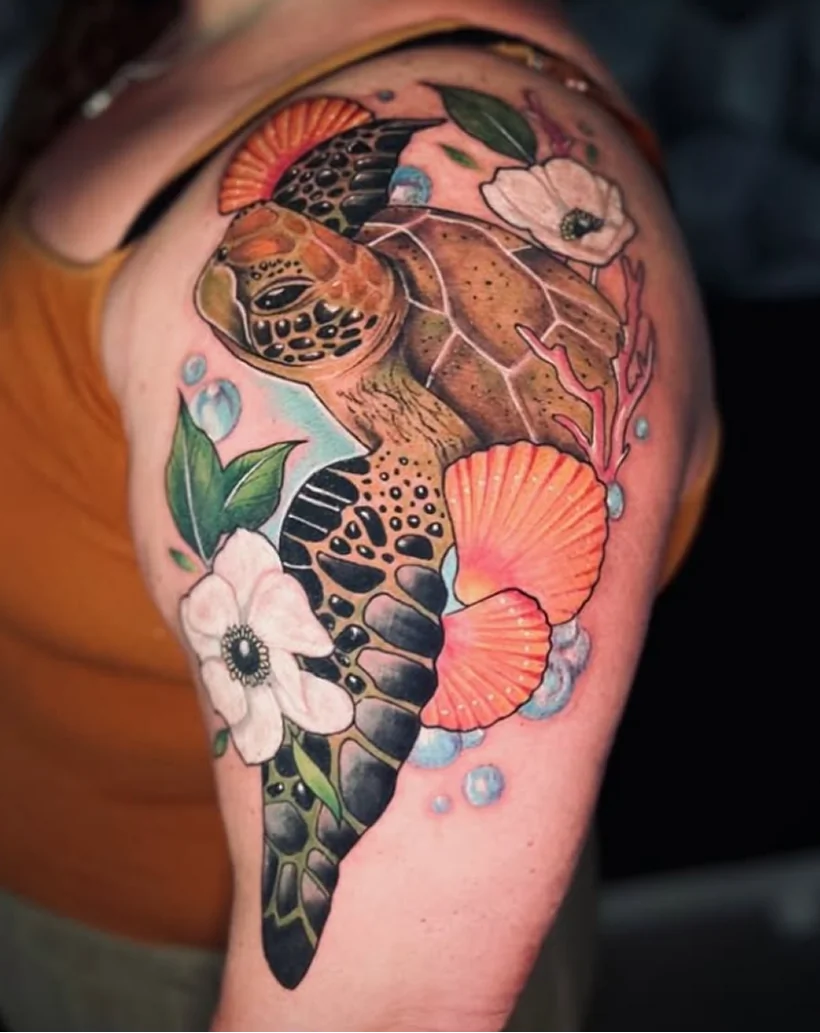 Color tattoo on an upper arm depicting a sea turtle swimming downward, surrounded by white flowers, pink seashells, coral branches, and blue bubbles