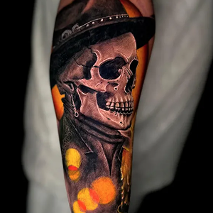 Color realism tattoo on an arm featuring a skull wearing a hat and dark jacket, highlighted by glowing orange and yellow light effects.
