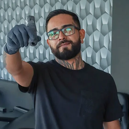 Portrait of tattoo artist David Ubaldo wearing glasses and black gloves, pointing a wrapped tattoo machine toward the camera.