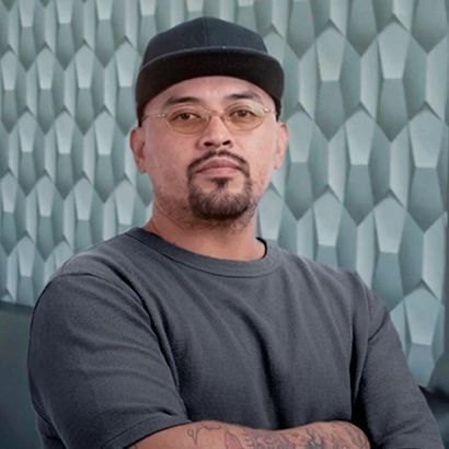 Portrait of tattoo artist Hammer with crossed arms, wearing a dark cap and tinted glasses against a grey geometric wall.