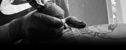 Close-up, black and white shot of a tattoo artist using a rotary machine to ink a design on a client's arm. Click to view our pricing.