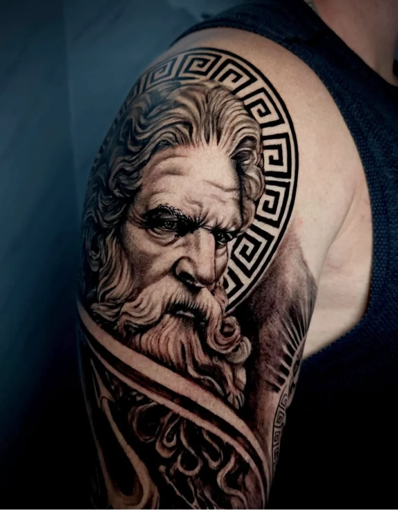 A highly detailed black and grey realism tattoo on a person's upper arm. It features a portrait of a stern, classical bearded man resembling a Greek god, with a circular Greek key pattern forming a halo behind his head.